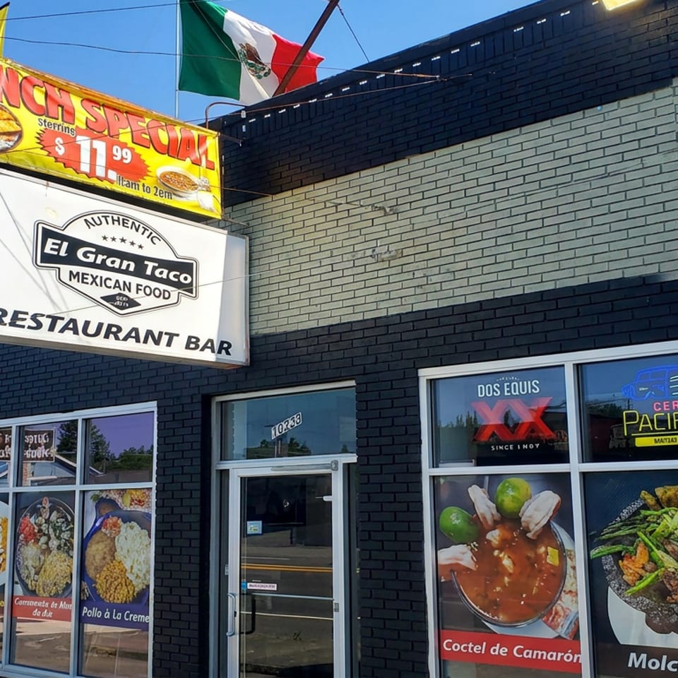 Exterior of El Gran Taco Mexican Food restaurant with lunch special sign, Mexican flag, and colorful food images in windows.