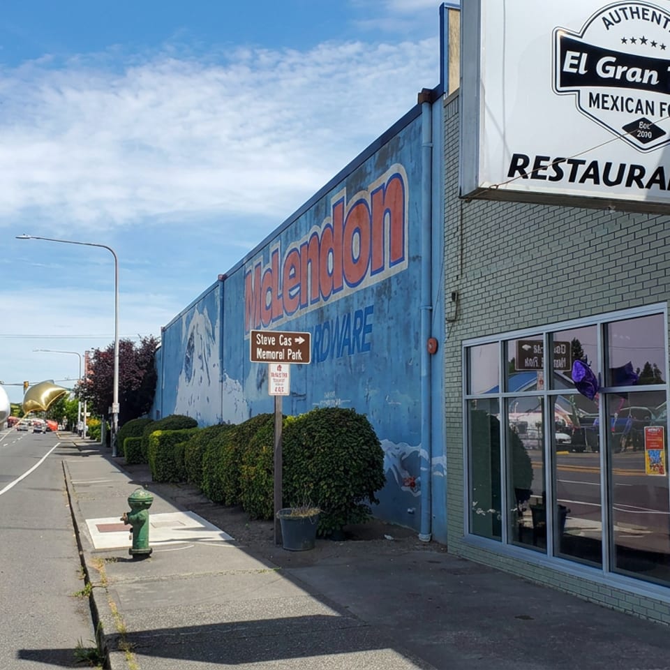 Street view of El Gran Taco Mexican restaurant with colorful signage, mural, sidewalk seating, and a sunny urban roadway.
